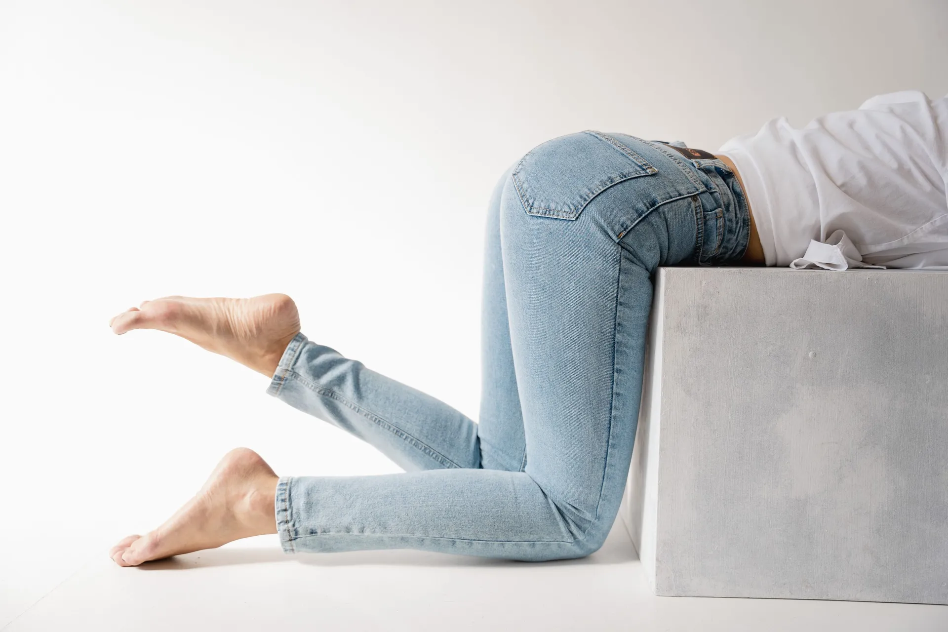 A model in light-wash skinny jeans kneels against a concrete block in a white studio