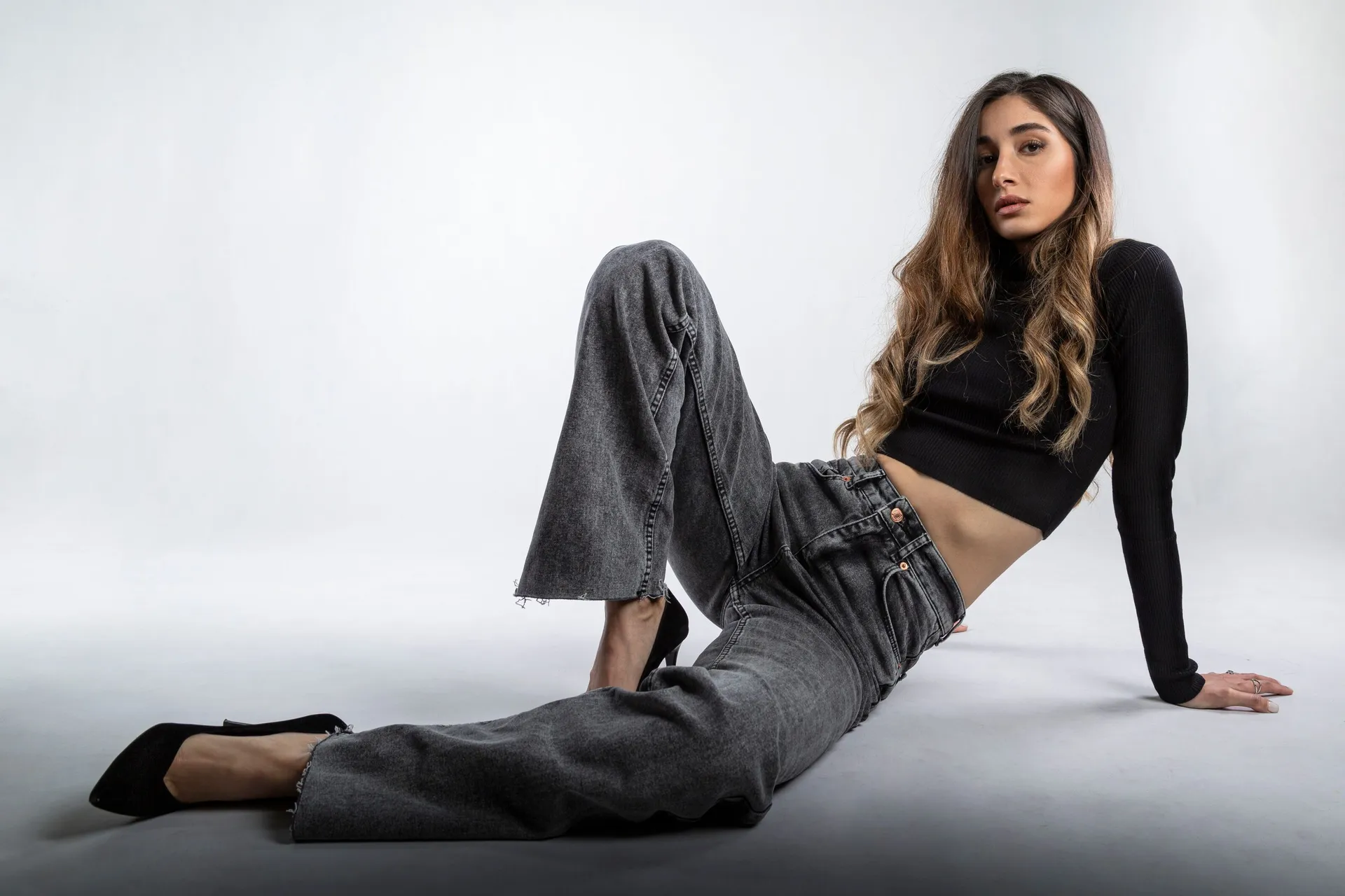 A model in grey wide-leg jeans and a black crop top reclines on a pale studio floor