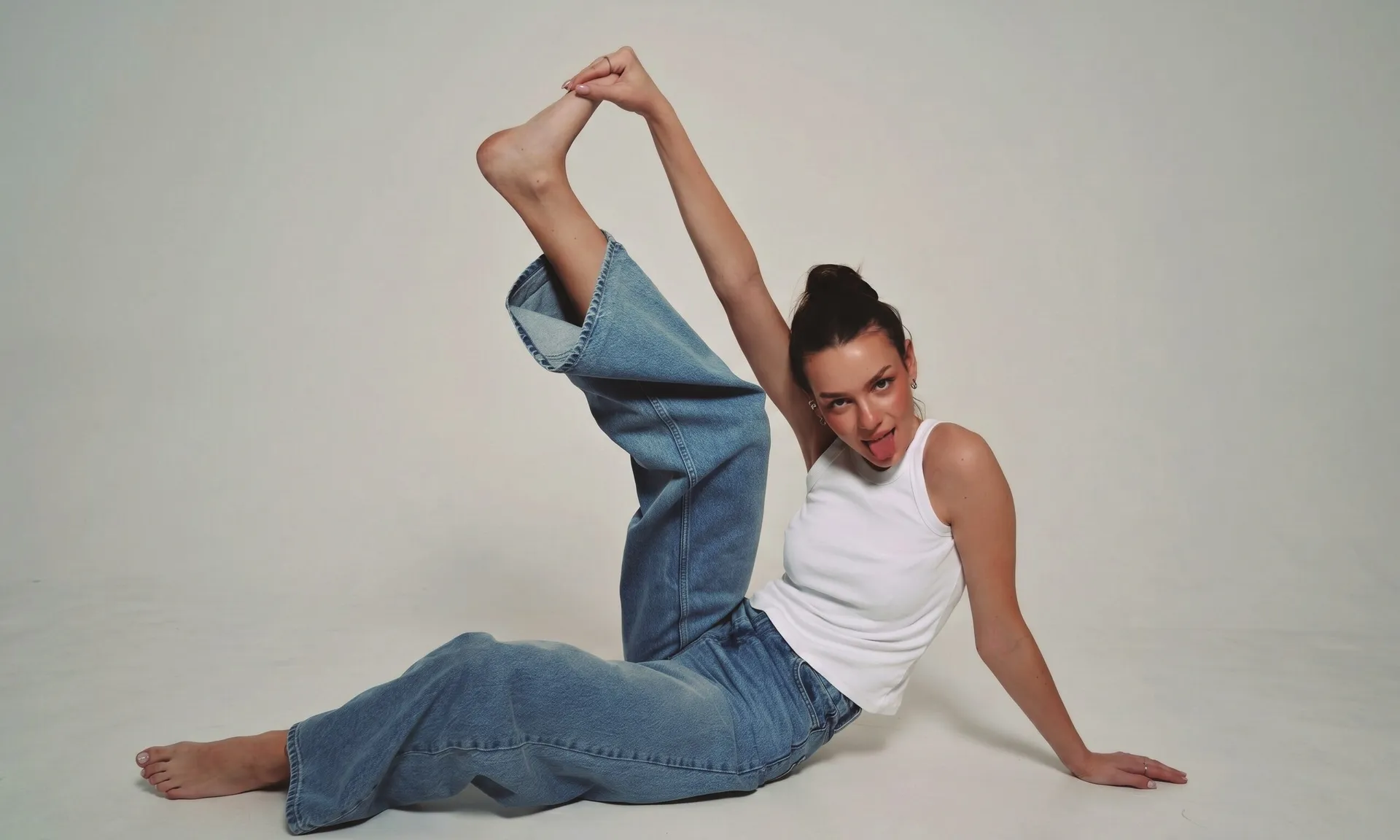 A model sits on the studio floor in light-wash wide-leg jeans, one leg raised high